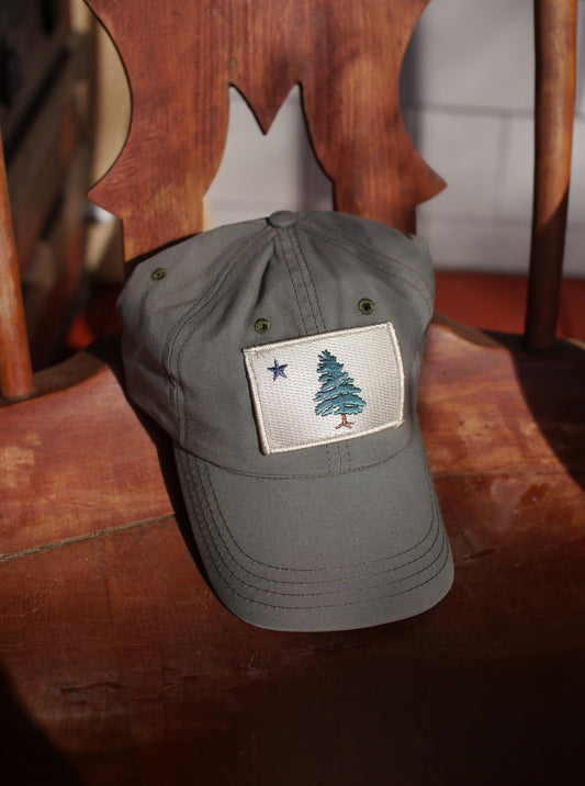 1901 Original Maine Flag insect-repelling baseball Cap in olive green sitting on a wooden chair in the sun.