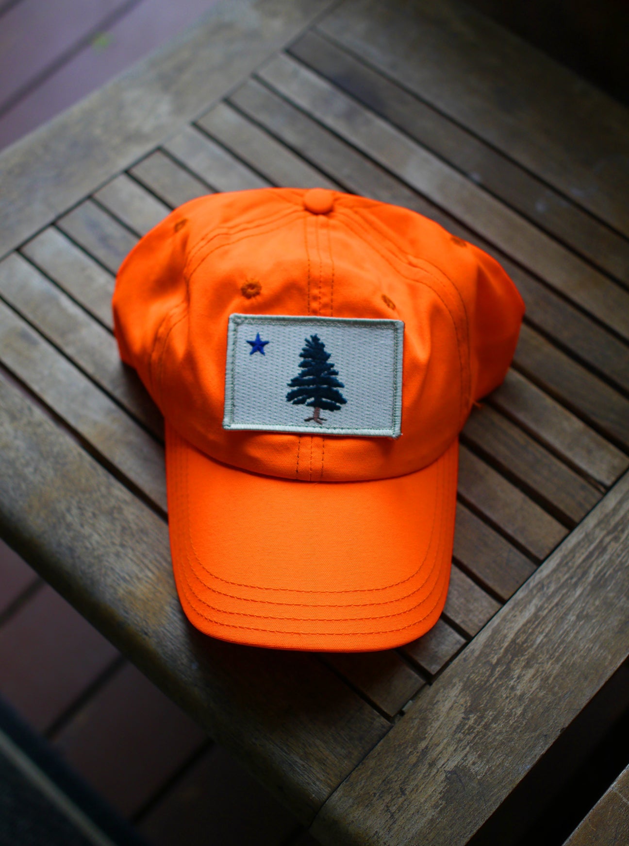 1901 Original Maine Flag insect-repelling baseball cap in hunter safe blaze orange sitting on a wooden bench in the sun.