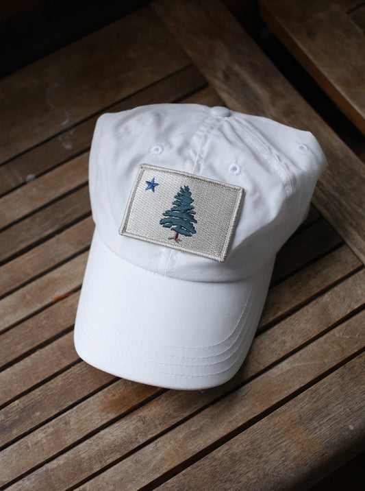 1901 Original Maine Flag insect-repelling baseball Cap in white sitting on a wooden chair in the sun.