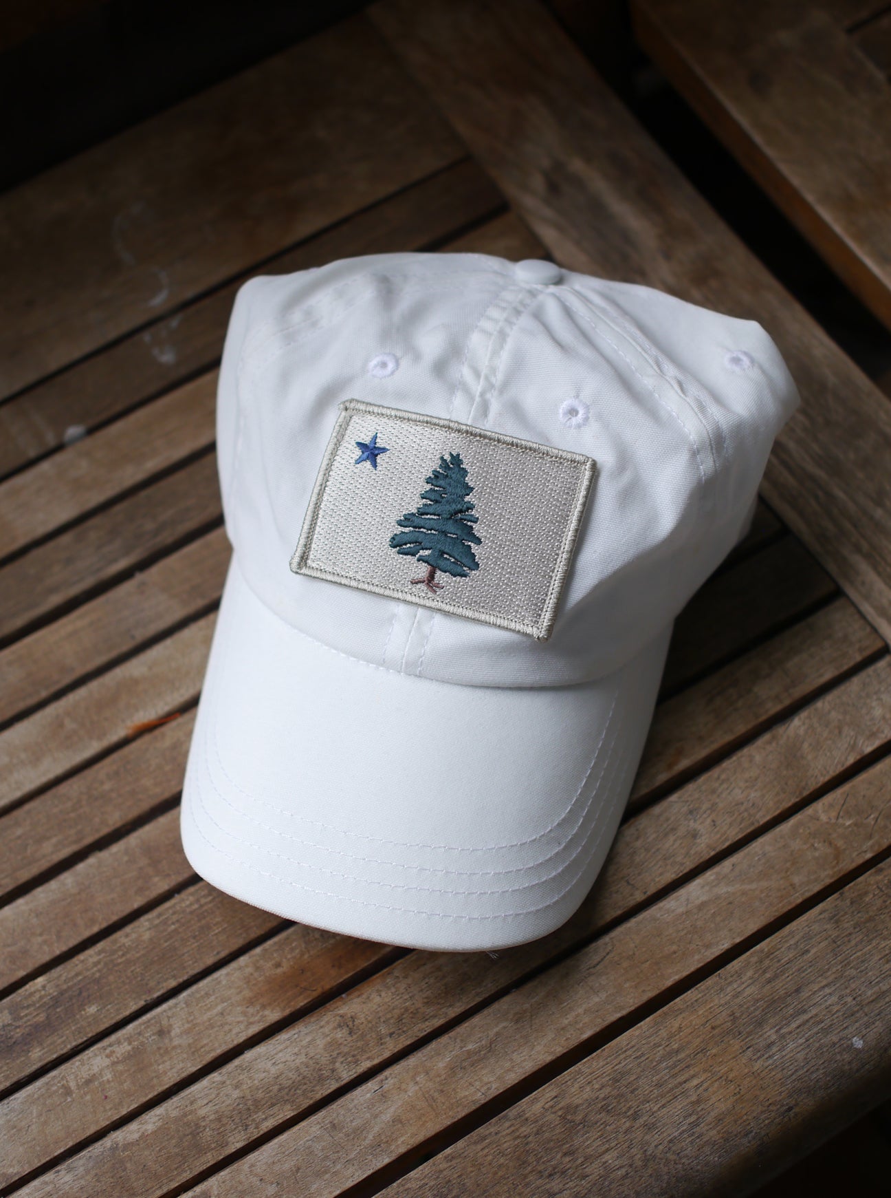 1901 Original Maine Flag insect-repelling baseball Cap in white sitting on a wooden chair in the sun. 